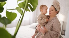 Young Mom With Baby Daughter In Her Arms In Bedroom With Green Plant In The Foreground, Mother Soothes Her Child With Pacifier In Mouth