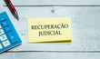 O texto Recuperação judicial em Português do Brasil escrito em um pedaço de papel que está sobre uma mesa de madeira. Uma calculadora e uma caneta na composição. Economia brasileira.