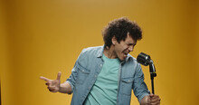 Close Up Shot Of Asian Man With Curly Hair In Casual Clothes Crazily Dancing And Singing In Microphone, Isolated On Yellow Background 
