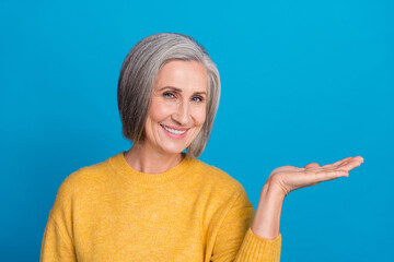 Poster - Photo portrait of senior gorgeous woman palm hold empty space product dressed stylish yellow garment isolated on blue color background