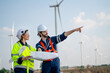 © narong - Caucasian engineer man point to right side and discuss together with co-worker woman hold drawing paper and stay in front of row of windmill or wind turbine with blue sky.