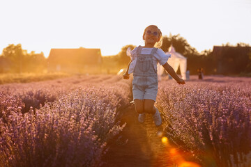 Wall Mural - adorable child girl in lavender field on sunset. smiling kid in sunglasses, jeans jumpsuit is having fun on nature on summer day. Family day, vacation, holiday. Girl up her hand