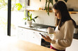 © Wavebreak Media - Non-binary trans woman holding a coffee cup using smartphone in the kitchen at home