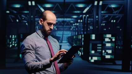 Poster - System administrator activating hardware in data center with tablet, analyzing modern servers in server room. Male infrastructure specialist looking at mainframe system. Tripod shot.
