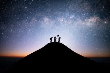 Wall Mural - Together overcoming obstacles as a group of three people raising hands up and holding flag on the top of a mountain. Celebrate victory and success over night sky background. Goal achievement symbol.