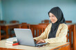 © tri mintarjo - beautiful asian young woman wearing hijab smiling at work, typing operating laptop computer and mobile phone at her desk.