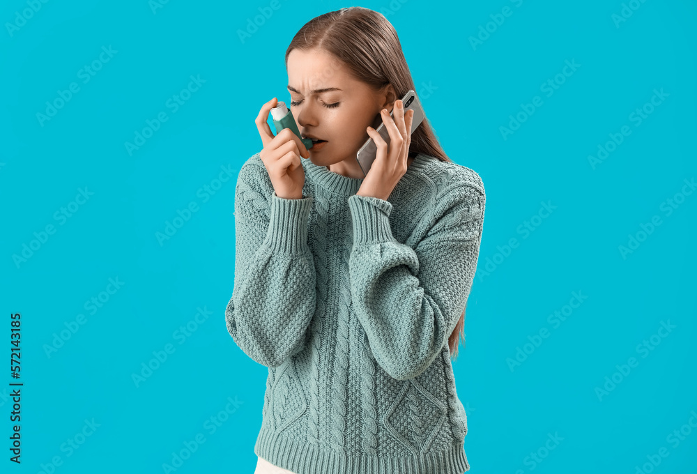 Sick young woman with inhaler calling an ambulance on blue background
