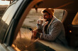 © La Famiglia - A handsome man in a denim jacket is sitting in the car truck, browsing a phone and drinking a hot drink in nature during the sunset.