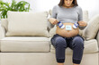 © Andrii - Cropped photo of pregnant woman sitting on the sofa with child clothes and paper on naked stomach.