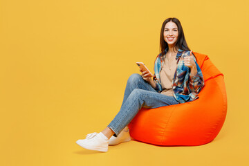 Wall Mural - Full body young fun woman wear blue shirt beige t-shirt sit in bag chair hold in hand use mobile cell phone show thumb up isolated on plain yellow background studio portrait. People lifestyle concept.