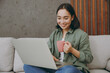 © ViDi Studio - Young fun IT woman of Asian ethnicity wear casual clothes work hold use laptop pc computer drink coffee sit on grey sofa couch stay at home hotel flat rest relax spend free spare time in living room.