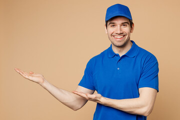 Poster - Happy delivery guy employee man wearing blue cap t-shirt uniform workwear work as dealer courier pointing hands arms aside indicate on area isolated on plain light beige background. Service concept.