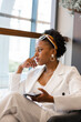 © Елена Бабанова - Pensive African American millennial woman with afro hairstyle in white suit wears yellow headband, sits in a cafe, relaxes, thinks and looks out the window. Black girl student takes a break from work