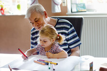 Cute Little Baby Toddler Girl And Handsome Senior Grandfather Painting With Colorful Pencils At Home. Grandchild And Man Having Fun Together