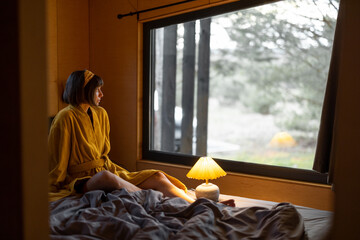 Wall Mural - Woman in yellow bathrobe wakes up in tiny bedroom of a wooden cabin in forest. Good morning and rest in house on nature concept