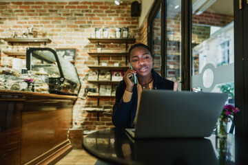 Wall Mural - Portrait of a smiling young woman using laptop and talking on mobile phone in coffeeshop