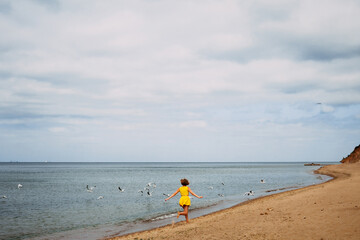 Little cute girl in yellow summer clothes runs after the birds on the seashore on a sunny day with clouds