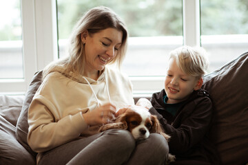 Portrait of happy beautiful middle-aged woman sitting on grey sofa with teenage boy at home, holding dog, stroking pet.