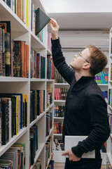 young adult man stands in library in middle of shelves and takes book. reading and recreation, educa