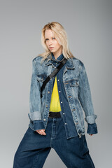 young blonde woman in stylish denim jacket posing while standing isolated on grey.