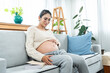 Beautiful Asian woman sitting on the sofa in the living room touching the belly. single mother who is pregnant puts down the book you are reading because of the onset of abdominal pain near delivery.