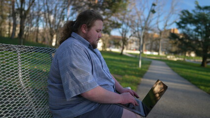 Young man using laptop on campus bench. Millennial guy working or studying remotely sitting outdoors in nature using modern technology