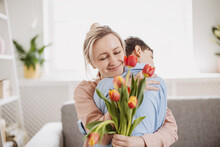 Cute Boy Sitting On The Sofa With Mom And Giving A Bouquet Of Tulips To Her.
