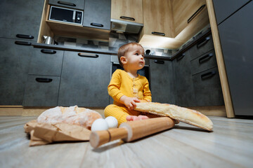 A cute one-year-old boy is sitting in the kitchen and eating a long bread or baguette in the kitchen. The first eating of bread by a child. Bread is good for children