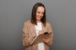 © sementsova321 - Photo of satisfied delighted woman with brown hair wearing beige jacket posing isolated over gray background, businesswoman writing to do list for working day.