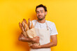 Food concept. Insecure handsome man baker in apron keeps bunch of fresh baked baguettes looks displeased and upset isolated over yellow wall