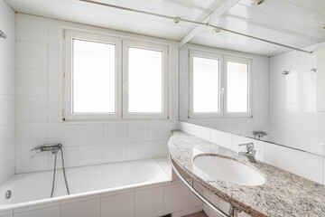 White bathroom with large hot tub. On the wall are two plastic windows with frosted glass through which daylight shines. Nearby is a sink on a marble countertop with a large mirror on the wall.