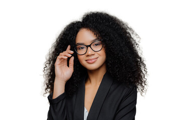 Close up portrait of calm satisfied Afro American woman with bushy curly hairstyle, keeps hand raised, has healthy skin, wears spectacles, black elegant outfit isolated on brown wall. Face expressions