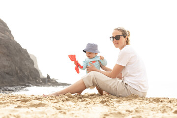 Wall Mural - Mother playing his infant baby boy son on sandy beach enjoying summer vacationson on Lanzarote island, Spain. Family travel and vacations concept