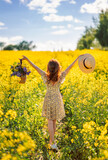 Fototapeta Kwiaty - girl with flowers and a hat in a field of rapeseed. Yellow flower field. View from the back