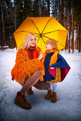 Wall Mural - Family with daughter and mother in orange dress with yellow umbrella having fun and joy in woods, forest or park with snow at winter day. Partial focus