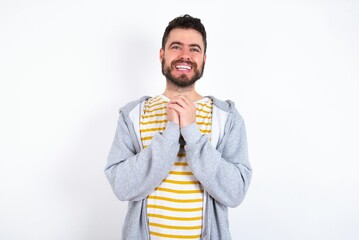 Wall Mural - Positive caucasian man wearing casual sportswear over white wall smiles happily, glad to receive pleasant news from interlocutor, keeps hands together. People emotions concept.