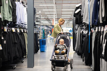 Casualy Dressed Mother Choosing Sporty Shoes And Clothes Products In Sports Department Of Supermarket Store With Her Infant Baby Boy Child In Stroller