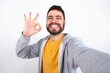 © Jihan - Portrait of pretty cheerful caucasian man wearing casual sportswear over white wall make selfie show okey symbol