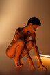 © LIGHTFIELD STUDIOS - full length of young and slim woman in lingerie sitting on haunches near fluorescent lamp on beige background.