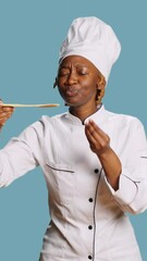 Wall Mural - Vertical video: African american cook doing food taste test with spoon, sipping soup for taste testing on camera. Young gourmet chef in apron tasting restaurant meal over blue background, tasty food.