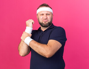 Wall Mural - aching adult slavic sporty man wearing headband and wristbands holding his hand isolated on pink background with copy space