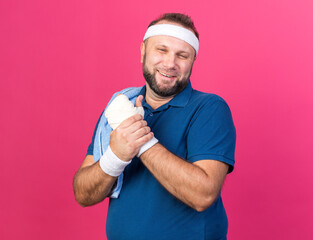 Wall Mural - smiling adult slavic sporty man with towel on shoulder wearing headband and wristbands holding his hand isolated on pink background with copy space