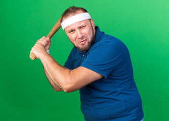 Wall Mural - scared adult slavic sporty man wearing headband and wristbands holding bat isolated on green background with copy space