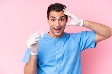 Wall Mural - Dentist caucasian man holding invisible braces isolated on pink background with surprise expression