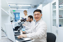 Portrait Of Asian Lab Technician, Man With Team Group Of People Working In Laboratory Researching, Man With Microscope In White Coat And Goggles Smiling And Looking At Camera.