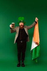 Wall Mural - Excited bearded man in hat holding glass of beer and Irish flag on green background.