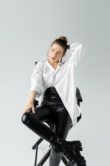 Wall Mural - stylish woman in black tight pants and white shirt holding hand behind head while sitting on chair on grey background.