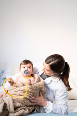 pediatrician playing with baby and stethoscope, smiling young woman in gown with hair up, blond Caucasian baby covered by brown blanket, with pacifier, looking at doctor