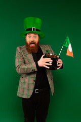 Wall Mural - Shocked bearded man in hat holding pot with coins and Irish flag isolated on green.