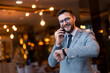 © Dorde - Smiling handsome businessman in gray silver suit and eyeglasses talking on smartphone, looking at hand watch, checking time, standing in modern restaurant waiting someone to arrive, expecting company.